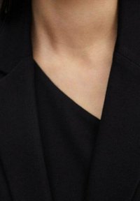 Veste noire avec un col structuré et un textile lisse. L'encolure présente une légère forme en V, mettant en avant des lignes épurées et un design moderne.