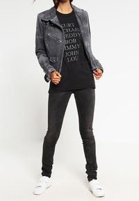 Schwarze Kunstleder-Motorradjacke mit silbernem Zubehör, ein grafisches schwarzes T-Shirt mit weißem Text und enge schwarze Jeans. Weiße Sneaker.