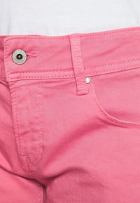Vaqueros de mezclilla rosa con cierre de botón plateado, costuras en los bolsillos frontales y un ligero brillo en la textura de la tela. Etiqueta de la marca visible.