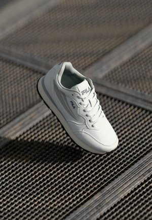 Hvit atletisk sneaker med overdel i lær, snøring, logodetalj og strukturert gummisåle, presentert mot et mønstret underlag.