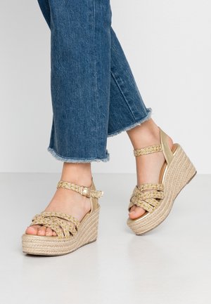 Pieds portant des sandales compensées à brides tressées beiges associées à un jean bleu coupé avec des bords effilochés sur un fond clair.