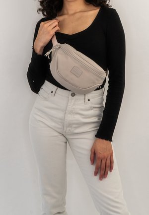 Osoba s kudrnatými vlasy, oblékající černý top s dlouhým rukávem a světlé kalhoty, přes rameno má béžovou kabelku přes tělo.