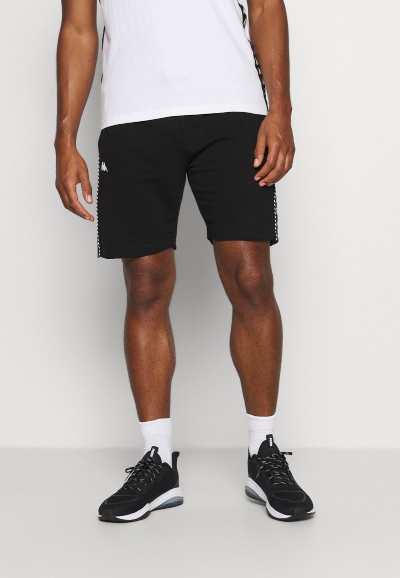 Shorts de sport noirs avec des bandes sur les côtés, fabriqués en matériau doux. Associés à des chaussures de course noires et des chaussettes blanches à la cheville.