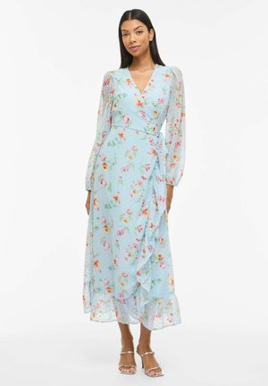 Moteris vilki šviesiai mėlyną, gėlėtą suknelę su permatomomis ilgomis rankovėmis ir raukšlėtu kraštu, derindama ją su sidabriniais dirželiais puoštus aukštakulnius, stovi baltame fone.