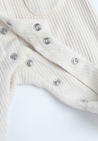 Vêtement en tissu côtelé crème avec des boutons-pression argentés le long de l'ouverture frontale et un encolure arrondie, mettant en valeur une texture douce.