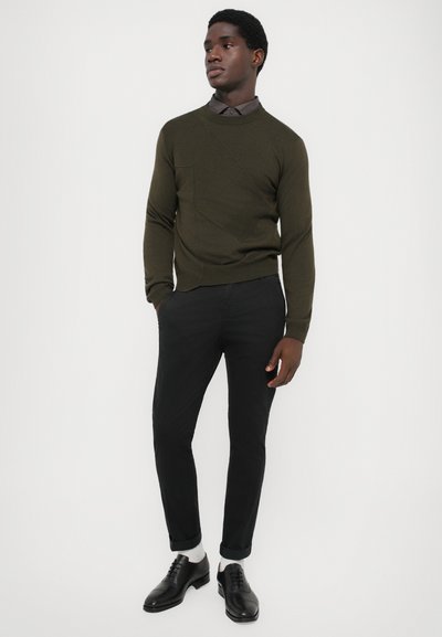 Maglione verde oliva con un motivo testurizzato, abbinato a pantaloni neri a sigaretta e scarpe eleganti nere. Sono visibili calzini grigio chiaro.