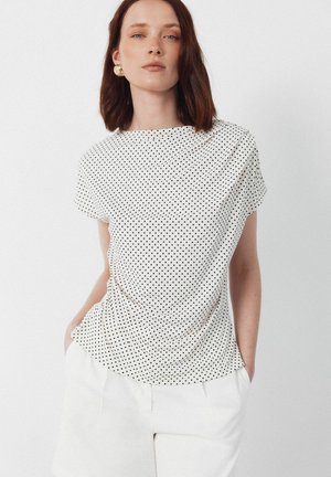Femme aux cheveux bruns mi-longs portant une chemise blanche à manches courtes avec des pois noirs et un pantalon blanc, les mains dans les poches.