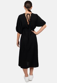 Vestito nero a lunghezza ginocchio in tessuto leggero e strutturato. Presenta una profonda scollatura a V sul retro, vita arricciata e maniche a sbuffo. Indossato con scarpe da ginnastica bianche.