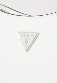 Sac à main blanc avec une texture brillante, portant un logo triangulaire argenté proéminent avec le texte "GUESS U.S.A." et un point d'interrogation.
