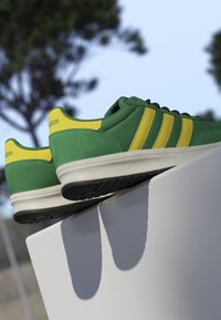 Pair of green and yellow Adidas sneakers with Cloudfoam soles resting on a white surface outdoors with blurred trees in the background.