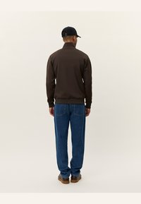 Καφέ μπουφάν με φερμουάρ και ψηλό γιακά, φορεμένο πάνω από μπλε τζιν με ίσια γραμμή. Μαύρο καπέλο ολοκληρώνει την εμφάνιση, διάσταση από πίσω.