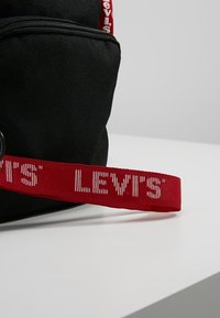 Zaino in tessuto nero con una tracolla rossa e il testo bianco "LEVI'S'", una tasca con zip e un dettaglio in metallo circolare.