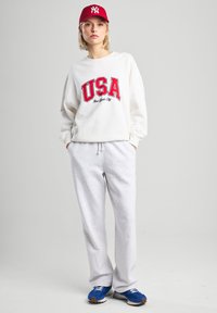 Felpa bianca con grafica rossa "USA" e testo "New York City", abbinata a pantaloni grigi e scarpe da ginnastica blu, completata da un berretto rosso.