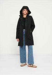 Black hooded parka with front snap closure, side pockets, and a relaxed fit. Worn over a black top and light blue flared jeans.