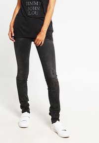 Schwarze, schmale Jeans mit einer abgetragenen Textur und dezentem Glanz, kombiniert mit weißen Sneakers und einem schwarzen T-Shirt mit gedrucktem Text.