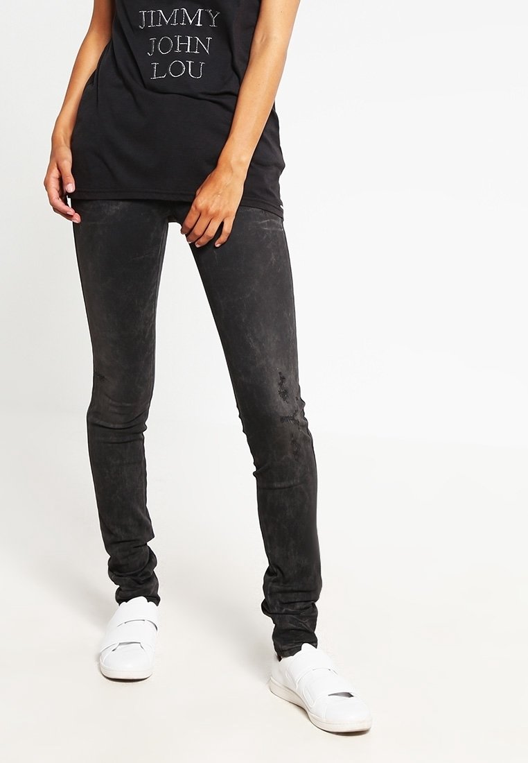 Schwarze, schmale Jeans mit einer abgetragenen Textur und dezentem Glanz, kombiniert mit weißen Sneakers und einem schwarzen T-Shirt mit gedrucktem Text.