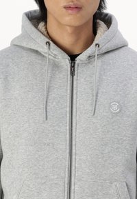 Grå lynlås-hoodie med fleeceforet hætte, der har snoretræk, sidelommer og et cirkulært logo-patch på brystet.
