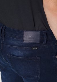 Pantalones vaqueros de mezclilla azul oscuro con una etiqueta de cuero que dice "BLEND". Incluyen bolsillos traseros y costuras en contraste sutil. Textura suave y diseño ajustado.