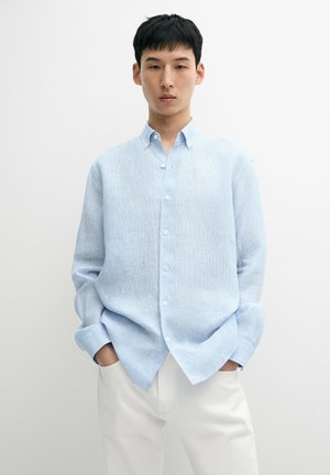 Junger Mann mit kurzen schwarzen Haaren, der ein hellblaues Hemd mit Knöpfen und weiße Hose trägt, die Hände in den Taschen vor einem schlichten Hintergrund.