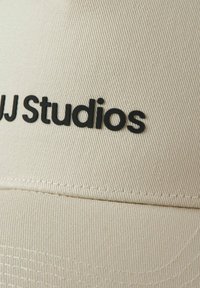 Beige cap with a textured fabric, featuring a raised black logo "J Studios" on the front. The stitching is visible along the brim.