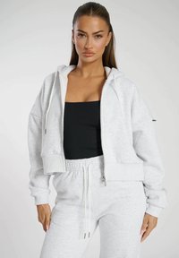 Felpa grigia corta in tessuto morbido, dotata di chiusura con zip, tasche frontali e cappuccio regolabile con coulisse. Abbinata a pantaloni coordinati.