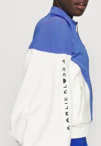 Blå og hvid lynlåstrøje med stribet øverste del, solid hvid nederste del og sort tekst på ærmet. Har tekstureret stof og høj krave.