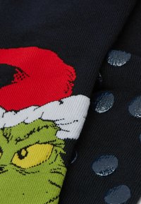 Zielona twarz Grincha z żółtymi oczami, nosząca czerwoną i białą czapkę Świętego Mikołaja na czarnym materiale z ciemnoniebieskimi, antypoślizgowymi kropkami.