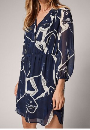 Frau trägt ein marineblaues V-Ausschnitt-Kleid mit abstraktem weißem und schwarzem Tiermuster und transparenten langen Ärmeln, steht vor einfachem Hintergrund.