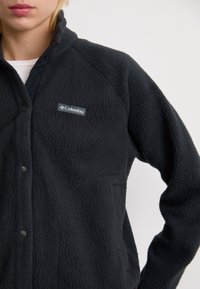 Close-up van een persoon die een zwarte Columbia fleecejas met drukknopen draagt, met het merklogo op de borst zichtbaar.