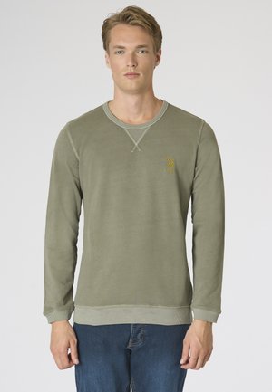 Giovane uomo in piedi, indossa una felpa verde oliva a maniche lunghe con un piccolo emblema giallo e jeans blu su sfondo uniforme.