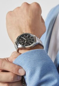 Montre en acier inoxydable argenté avec un cadran noir, sous-cadrans chronographe et un bracelet maillons, portée sur une chemise en tissu texturé bleu clair.