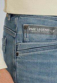 Light blue denim jeans featuring a dark gray label "PME LEGEND AMERICAN CLASSIC," with contrasting stitching and a buttoned pocket detail.
