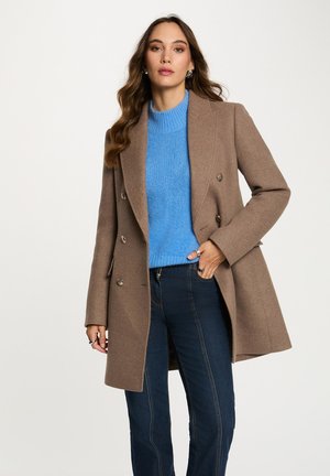 Manteau croisé en laine marron avec des boutons métalliques, associé à un pull en laine bleu et un pantalon en denim sombre avec des détails de couture visibles.