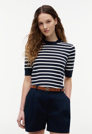 Νεαρή γυναίκα με σγουρά μαλλιά, φορώντας μπλε σκούρο και λευκό ριγέ κοντομάνικο τοπ, μπλε σκούρες βερμούδες και καφέ ζώνη με αγκράφα λογότυπο.