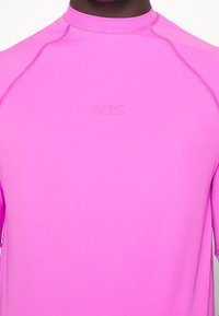 Gros plan d'une personne portant une chemise rose vif avec « GCDS » embossé sur la poitrine et des coutures visibles le long des épaules et des manches.