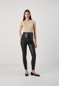 Beige ribbad utan ärm topp som matchas med högmidjade svarta fusk läder leggings och platta svarta skor. Slät textur, kroppsnära design.