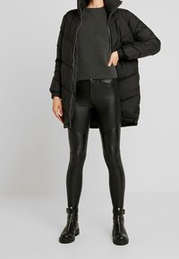 Svart puffjacka med hög krage, matchad med en mörkgrå texturerad tröja och svarta läderleggings med ribbade detaljer. Svarta stövlar fulländar looken.