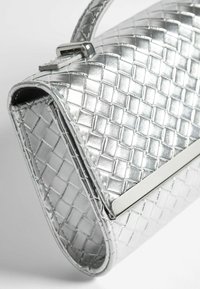 Borsa a mano tessuta in argento con un design strutturato, caratterizzata da una chiusura in metallo elegante e una finitura lucida, posizionata in diagonale su uno sfondo chiaro.