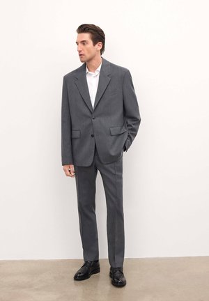 Traje gris con solapas de pico y dos bolsillos frontales, combinado con una camisa blanca y zapatos de vestir negros. Tejido suave y corte a medida.