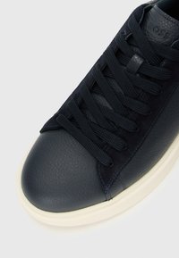 Sneaker din piele și velur bleumarin cu șireturi negre și talpă din cauciuc alb, arătând vârful texturat și logo-ul embosat "BOSS" pe limbă.