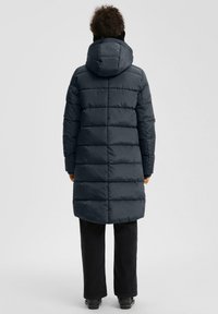 Žmogus, vilkintis ilgą tamsią pūkinę striukę su kapišonu, juodas kelnes ir juodus batus, stovi atsigręžęs į šviesų fono paviršių.