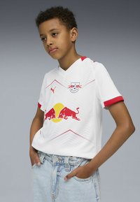Camiseta de fútbol blanca con detalles en rojo, que presenta un patrón en forma de chevron, un logo y dos toros rojos sobre un círculo amarillo. El modelo lleva pantalones cortos de mezclilla.