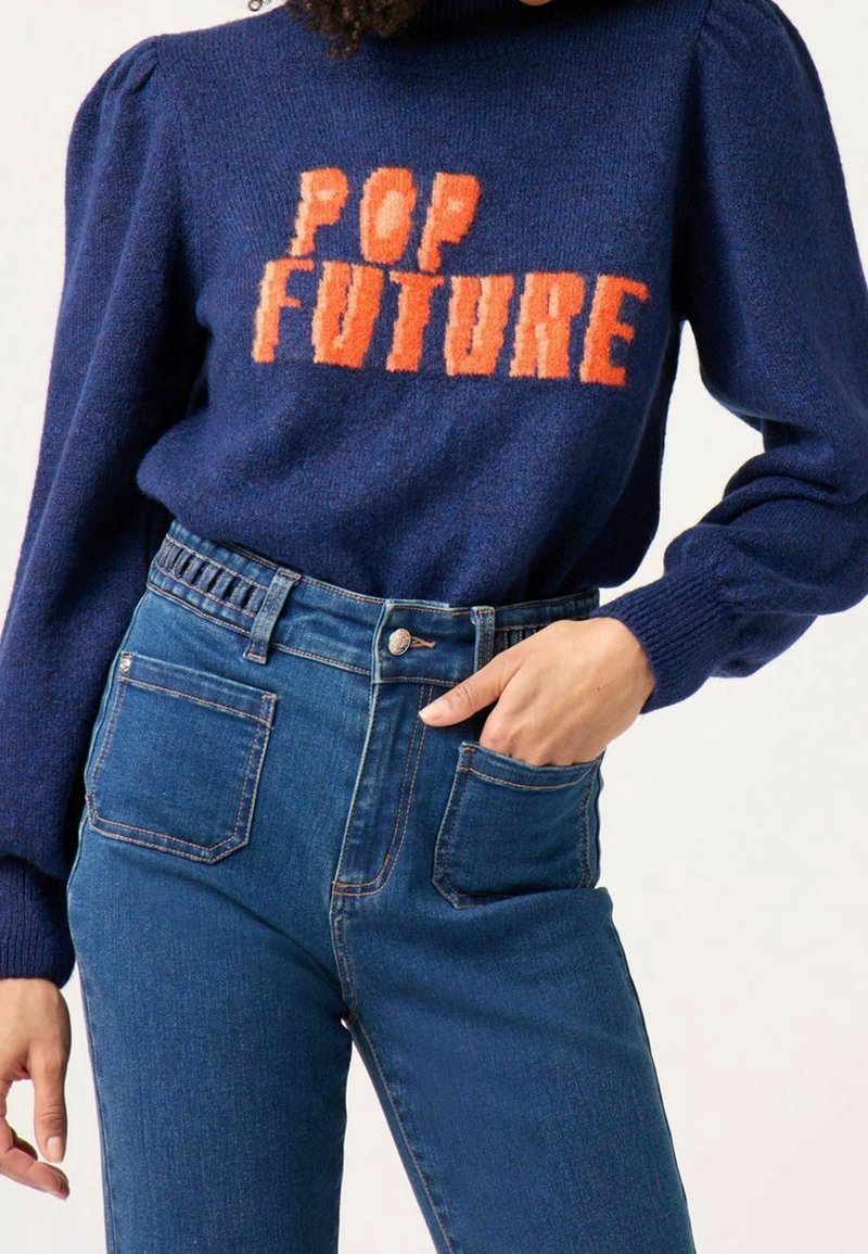 Pull en tricot bleu marine avec "POP FUTURE" en orange, avec des manches bouffantes et un col montant, associé à un jean bleu taille haute.