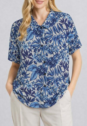 Donna che indossa una camicia a maniche corte floreale blu e bianca con bottoni infilata in pantaloni beige chiari, in piedi con le mani in tasca.