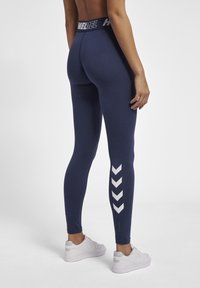I leggings blu navy presentano una ampia vita nera con branding, un motivo a chevron bianco sulla gamba sinistra e una texture aderente e liscia.