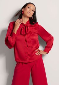 Rote Satinbluse mit einer Schlaufe am Kragen, Knopfverschluss vorne und langen Ärmeln mit gerafften Manschetten. Glatte Textur und lebendige Farbe.