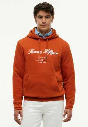 Junger Mann trägt einen orangefarbenen Tommy Hilfiger Hoodie über einem blauen Hemd mit Knöpfen und weißen Hosen, steht vor einem schlichten Hintergrund.