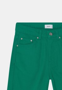 Gröna denimbyxor med framknapp, bältesöglor och nitar på framsidan och myntlommen, etikett med texten "GRUNT" på insidan av midjebandet.