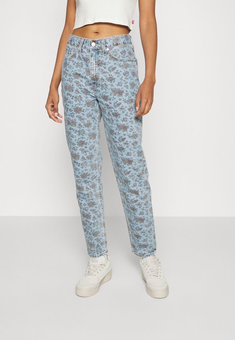 Jean bleu clair à motif floral avec une coupe droite, fermeture par bouton et design à cinq poches, associé à des baskets blanches.