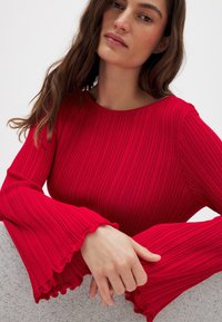 Rød ribbet striktrøje med rund halsudskæring, flagrende ærmer og flæsekanter. Glat tekstur og tætsiddende design, der fremhæver formen.